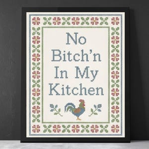 May include: Framed cross-stitch art with the text "No Bitch'n In My Kitchen." The design features a rooster and floral border in blue, green, and red on an off-white background. The artwork is displayed in a black frame.