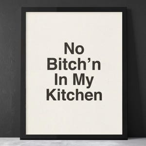 May include: Framed art print with the text "No Bitch'n In My Kitchen" in bold, black letters on an off-white background. The print is encased in a black frame, suitable for kitchen decor.