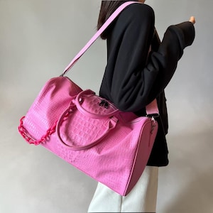 May include: A vibrant pink duffel bag with a crocodile skin effect. It features a matching pink shoulder strap and a pink chain detail. The bag is held by a person wearing a black top and white trousers.