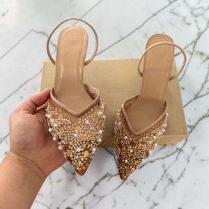 Puede incluir: Un par de tacones beige de punta fina con una delicada correa en el tobillo. La parte superior del zapato está adornada con lentejuelas y pequeñas cuentas redondas. Los tacones están sobre un fondo neutro.