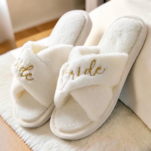 以下が含まれることがあります： 白くふわふわした花嫁用スリッパのペア。クロスデザインで、「Bride」の文字が各スリッパの上部に金色の筆記体で刺繍されています。スリッパは白く、質感のある表面に置かれています。