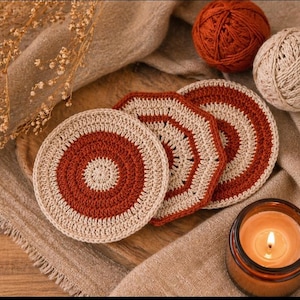 May include: Handmade crochet coasters in a warm color palette. The coasters feature a circular design with a rust-colored center and a cream-colored border. Two balls of yarn and a lit candle are also in the image.