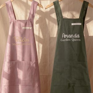 May include: Two aprons hanging on wooden hangers. The left apron is dusty pink with the embroidered text 'Felicia Mama's Kitchen'. The right apron is dark olive green with the embroidered text 'Amanda Garden Queen'. Both feature cross-back straps and two front pockets.