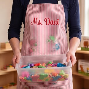 Könnte beinhalten: Eine rot-weiß vertikal gestreifte Schürze mit dem gestickten Text Ms. Dani in roter Schreibschrift. Die Schürze hat eine große Vordertasche und ist mit verschiedenen bunten Farbspritzern versehen. Sie wird über einem dunkelblauen Langarmshirt getragen.