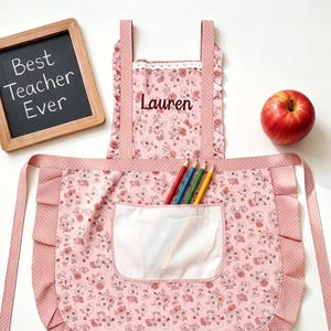 Op de afbeelding: Roze bloemen schort met een witte zak en ruches. De schort heeft de naam "Lauren" geborduurd en bevat kleurpotloden. Een klein schoolbord toont de tekst "Best Teacher Ever".