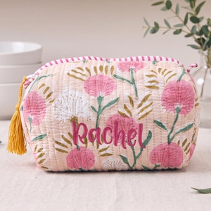 May include: A quilted fabric pouch in a light peach color featuring a floral pattern in shades of pink, white, and green. The name Rachel is embroidered in dark pink script on the front. A yellow tassel is attached to the side zipper pull.
