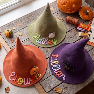 May include: Three pointed hats in olive green, orange, and purple. The olive hat features the name Hailey and a cat patch. The orange hat displays the name Elowen and a pumpkin patch. The purple hat shows the name Lillian and a bat patch.