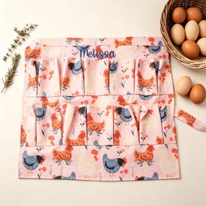 May include: A pink egg gathering apron with a repeating pattern of chickens in various colors. The apron has multiple pockets and is personalized with the name "Melissa" in blue script. A basket of eggs is in the background.