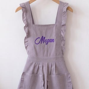 Puede incluir: Un delantal de color morado claro con finas rayas verticales blancas, con tirantes con volantes, un dobladillo con volantes y dos bolsillos delanteros. El nombre Megan está bordado en letra cursiva morada en el pecho del delantal.