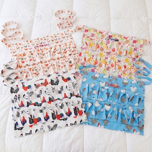 May include: Four colorful egg gathering aprons with ruffled pockets and tie straps. The aprons feature various patterns, including chickens, roosters, and Easter egg designs. Each apron has a name embroidered on it: Brianna, Jocelyn, Jennifer, and Natalie.