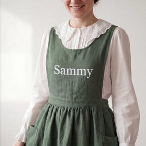 Könnte beinhalten: Eine olivgrüne Latzschürze mit dem in weißem Garn auf die Brust gestickten Namen Sammy. Die Schürze hat einen gerafften Rock mit zwei sichtbaren Taschen und wird über einer weißen langärmeligen Bluse mit einem gewellten Spitzenkragen getragen.