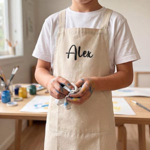 Könnte beinhalten: Eine beigefarbene Schürze aus Segeltuch mit dem Namen Alex, der in schwarzer Schreibschrift auf die Brust gestickt ist. Die Schürze hat breite Schulterträger und eine große Vordertasche und wird über einem weißen T-Shirt getragen.