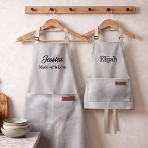 May include: Two gray and white pinstriped aprons hanging on wooden hangers. The larger apron features the embroidered text Jessica and Made with Love, while the smaller apron features the text Elijah. Both aprons have a front pocket with a brown label.