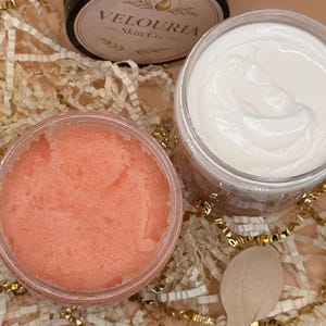 May include: An overhead shot of skincare products. The image features three open jars of products, including a pink scrub, a white cream, and a jar with a black lid. A wooden spoon rests near the jars. The products are on a bed of gold and white decorative paper.