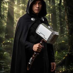 May include: A person wearing a black hooded cloak, holding a silver and brown hammer. The hammer has intricate detailing and a leather strap. The background is a lush forest with sunlight filtering through the trees.