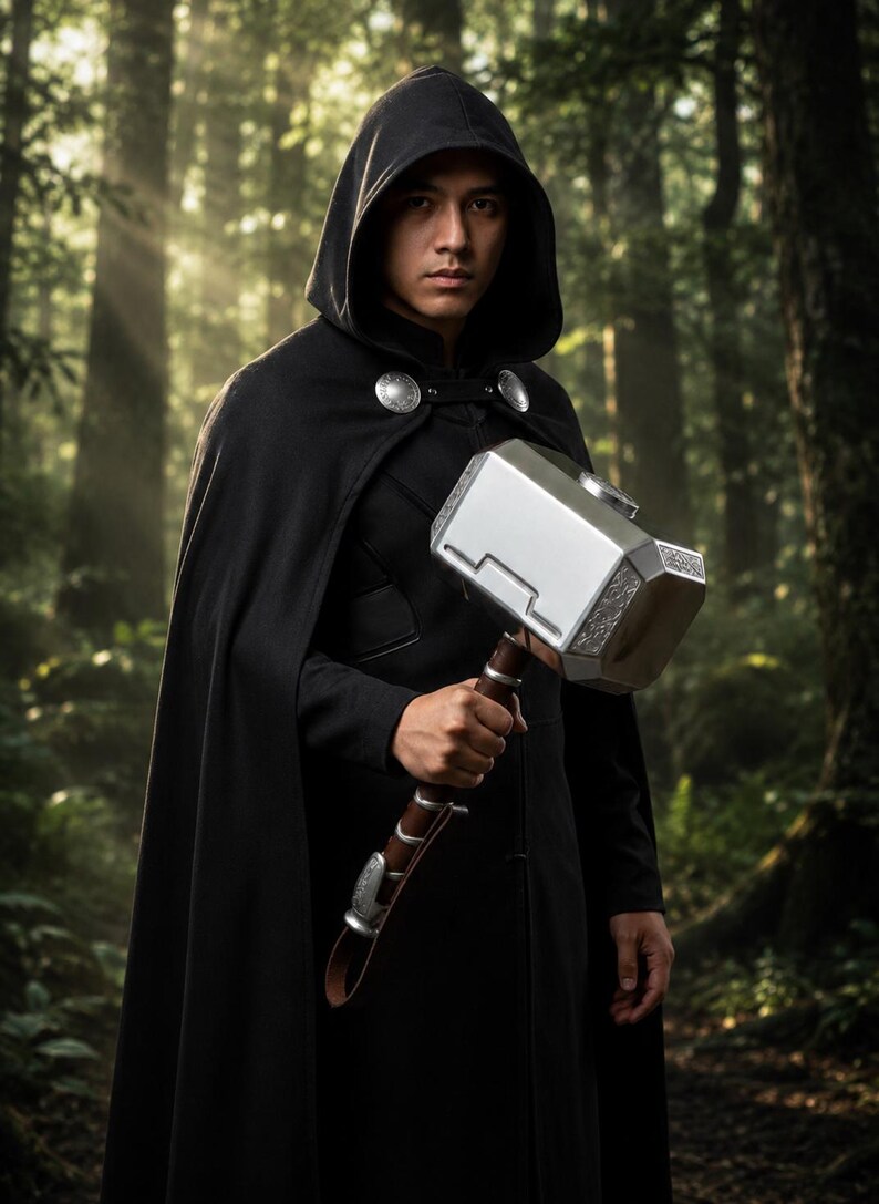 May include: A person wearing a black hooded cloak, holding a silver and brown hammer. The hammer has intricate detailing and a leather strap. The background is a lush forest with sunlight filtering through the trees.