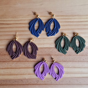 May include: A collection of handmade polymer clay earrings in various colors, including blue, purple, and green. Each pair features a unique, abstract design with gold-colored posts. The earrings are arranged on a wooden surface.