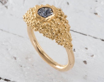 Könnte beinhalten: Ein goldener Ring mit einer strukturierten Band und einem kleinen, grauen, rautenförmigen Stein, der in der Mitte eingesetzt ist.