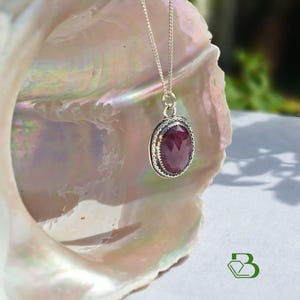 May include: A silver necklace featuring an oval-shaped, faceted deep red gemstone pendant. The pendant is set in a textured silver frame with a beaded edge and hangs from a delicate silver chain. The piece is displayed against a large, iridescent shell.