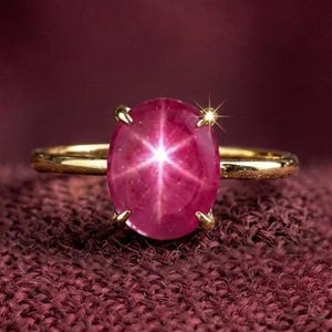 May include: A gold ring featuring a large, oval-shaped ruby gemstone with a six-ray star effect. The ruby is a deep pinkish-red color, set in a simple gold band. The ring is displayed on a textured, maroon fabric.