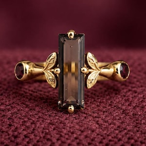 May include: A gold ring featuring a central rectangular baguette-cut smoky quartz stone flanked by two small round dark red garnet stones. The gold band is detailed with leaf-shaped metal accents on either side of the central gemstone setting.