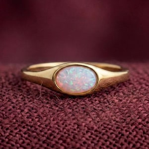 Può includere: Un anello d'oro con una pietra di opale ovale. L'opale presenta un mix di colori bianco, rosa e blu. L'anello è appoggiato su un tessuto rosso scuro.