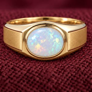 Peut inclure: Une bague en or avec une opale ovale centrale présentant des éclats irisés. L'anneau présente une finition dorée polie et lisse autour du sertissage, ainsi qu'une texture mate et brossée sur les côtés de la tige de la bague.