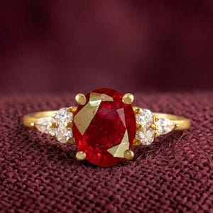 May include: A gold ring featuring a large, oval-cut, deep red gemstone at the centre, held by four prongs. The band is accented on each side by a cluster of three small, clear, round-cut white stones, creating a symmetrical and elegant design.