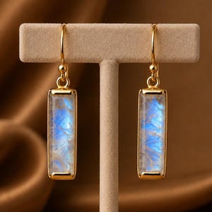 Könnte beinhalten: Ein Paar goldfarbene Ohrhänger mit rechteckigen, durchscheinenden, mondsteinartigen Edelsteinen, die einen bläulichen, irisierenden Schimmer aufweisen. Die Steine sind in einen glatten Goldmetallrahmen gefasst und hängen an einfachen Ohrhaken.