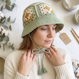 May include: A crocheted bucket hat in shades of green, cream, and yellow. The hat features a square pattern on the crown and a brim. It has a chin strap with tassels. A small tag is attached to the hat.