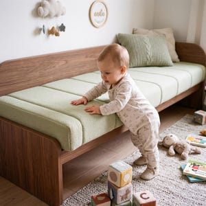 Puede incluir: Un diván de madera de perfil bajo con un cojín tapizado en verde claro con acolchado vertical. La estructura está fabricada en madera de tonos cálidos con veta visible, proporcionando una base sólida para el cojín texturizado.
