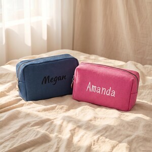 Puede incluir: Dos neceseres personalizados. Uno es azul marino con el nombre "Megan" bordado en negro. El otro es rosa con el nombre "Amanda" bordado en blanco. Ambos neceseres tienen forma rectangular y cierre de cremallera.