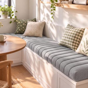 May include: A long, light gray bench cushion with a textured, channel-tufted surface. The cushion sits atop a white built-in storage bench and is accented by several square throw pillows in green and white floral, gingham, and botanical patterns.