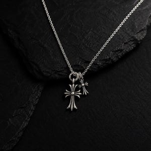 Op de afbeelding: Een zilveren ketting met twee kruisvormige hangers. Het grotere kruis heeft een gedetailleerd, sierlijk ontwerp, terwijl het kleinere kruis eenvoudiger is. De ketting is delicaat en de hangers hangen tegen een donkere achtergrond.