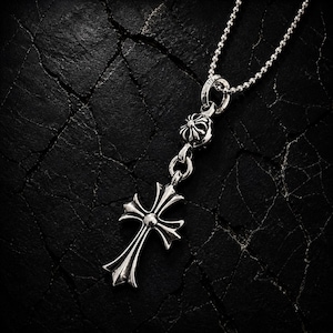 May include: A silver pendant necklace featuring a detailed cross design. The cross has ornate, pointed ends and a central sphere. The necklace includes a small floral accent and a delicate chain. The jewelry is displayed against a dark, textured background.