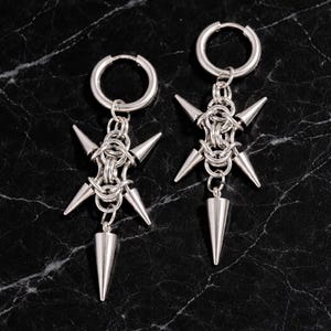 Pode incluir: Um par de brincos prateados com uma estética punk rock. Cada brinco apresenta um aro, um centro em estilo cota de malha e múltiplas pontas em forma de cone. Um único cone maior pende da parte inferior de cada brinco.