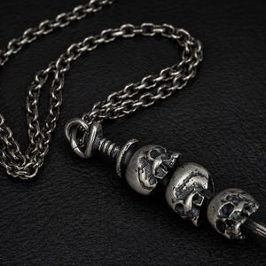May include: A silver-toned necklace featuring a chain and a pendant with three skull charms. The skulls are detailed with visible facial features and are attached to a central bar. The necklace is displayed against a dark background.