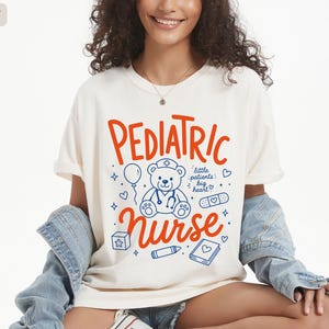 May include: An ivory short-sleeve t-shirt featuring the orange text PEDIATRIC NURSE and a blue graphic of a teddy bear wearing a stethoscope, surrounded by a balloon, a bandage, a block, a crayon, a book, and the text little patients big heart.