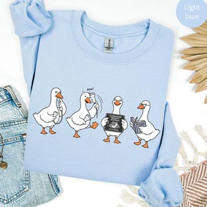 Op de afbeelding: Een lichtblauw sweatshirt met ronde hals en een grafische print van vier witte ganzen. De ganzen zijn afgebeeld tijdens verschillende activiteiten, waaronder het vasthouden van een echoscopie-sonde, het slaan op een sonde, het vasthouden van een bord met de tekst ULTRASOUND RESULTS CONFIRMED IT'S A GOOSE en het lezen van een boek.