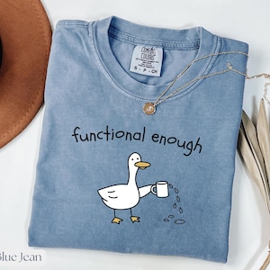 Könnte beinhalten: Blaues Jeans-T-Shirt mit dem Schriftzug "functional enough" und einer Cartoon-Ente, die eine Kaffeetasse hält. Das Shirt hat einen Rundhalsausschnitt und kurze Ärmel. Eine goldene Halskette mit Anhänger liegt auf dem Shirt.
