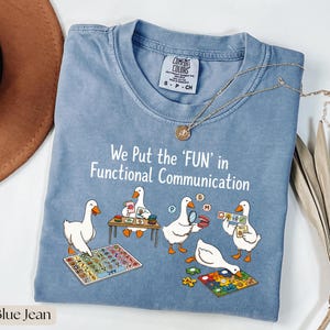 Könnte beinhalten: Ein blaues T-Shirt mit einer Grafik von fünf weißen Gänsen, die mit Kommunikationshilfen interagieren. Der Text lautet, We Put the 'FUN' in Functional Communication. Das T-Shirt hat einen Rundhalsausschnitt und ein Comfort Colors Etikett innen.
