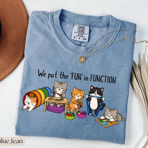May include: A blue jean colored t-shirt featuring a graphic of five cartoon cats playing with various therapy tools. The text "We put the 'FUN' in FUNCTION" is printed in black above the cats. The shirt has a Comfort Colors brand label.