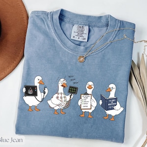 Peut inclure: T-shirt bleu jean avec un motif fantaisiste de quatre oies de dessin animé dans des rôles médicaux. Les oies tiennent des appareils médicaux et des livres. Le t-shirt porte le texte "COMFORT COLORS" et "S P CH". Un collier en or est drapé sur le t-shirt.