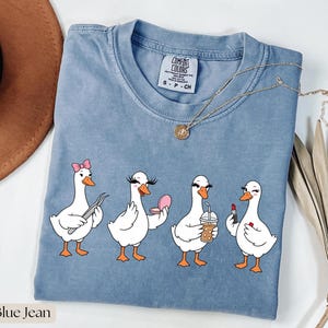 Op de afbeelding: Een blauw spijkerkleurig t-shirt met een afbeelding van vier witte ganzen. De eerste gans draagt een roze strik, de tweede houdt een compact spiegeltje vast, de derde drinkt uit een plastic beker met een rietje en de vierde brengt rode lippenstift aan.