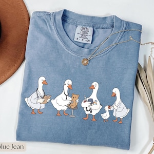 Puede incluir: Una camiseta de color azul vaquero con un gráfico de cuatro gansos blancos vestidos como profesionales médicos. Los gansos aparecen realizando diversas tareas, como escribir en un portapapeles, usar un estetoscopio en un osito y examinar a un gansito.