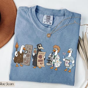 Pode incluir: Camiseta azul jeans com uma ilustração de desenho animado de gansos vestidos como figuras históricas e profissionais médicos. Os gansos estão segurando vários itens, incluindo uma tocha, uma lupa e um microscópio. A camisa tem o texto "Comfort Colors".
