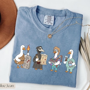Op de afbeelding: Blauw Comfort Colors T-shirt met een cartoonontwerp van vier ganzen in verschillende beroepen. De ganzen zijn gekleed als een holbewoner, een pestdokter, een verpleegster en een barista. Een gouden ketting is over het shirt gedrapeerd.