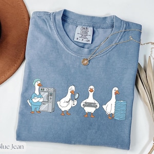 May include: Blue jean coloured Comfort Colors t-shirt with a crew neck. The shirt features a cartoon design of four white geese in medical attire, each holding different medical tools. A gold necklace with a pendant is draped across the shirt.