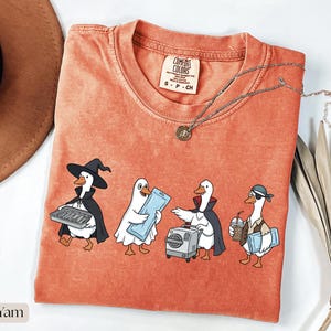 May include: A burnt orange short-sleeve t-shirt featuring a graphic of four cartoon ducks dressed in various costumes, including a witch, a ghost, a vampire, and a pirate. The shirt has a crew neckline and a Comfort Colors brand label inside.