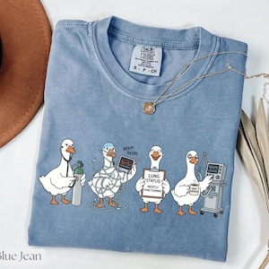 May include: Blue jean colored Comfort Colors t-shirt with a humorous graphic of cartoon geese in medical scenarios. One goose holds an oxygen tank, another is hooked up to monitors, and another holds a sign that says "LUNG STATUS: MOSTLY FUNCTIONING."